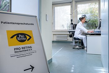 Aufsteller einer PRO RETINA-Sprechstunde Ein Aufsteller mit PRO RETINA-Logo und Pfeil zeigt auf eine Person, die an einem Schreibtisch sitzt.